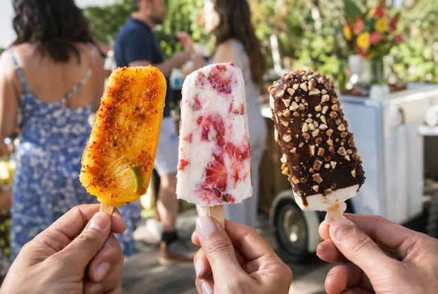 Three hands hold a mango chile paleta, a strawberries and cream paleta and a chocolate-dipped coconut paleta.